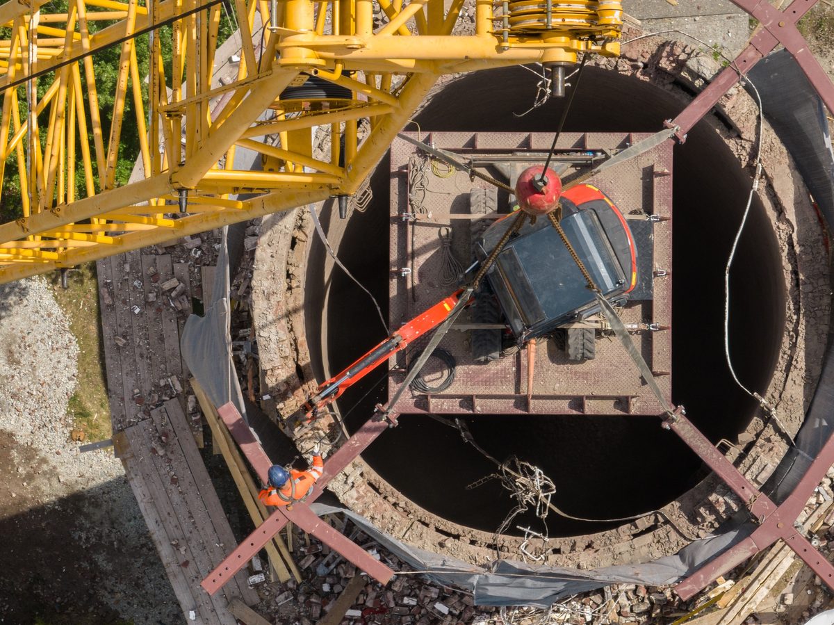 Aerial view of a crane lowering a machine on a steel platform into the top of a brick chimney.