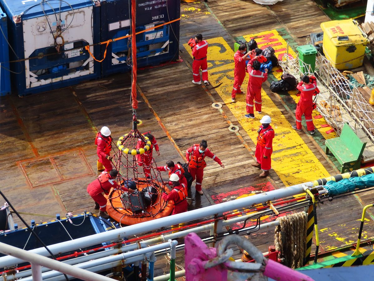 Offshore crew in red coveralls loading bags into a personnel transfer basket on a platform deck.
